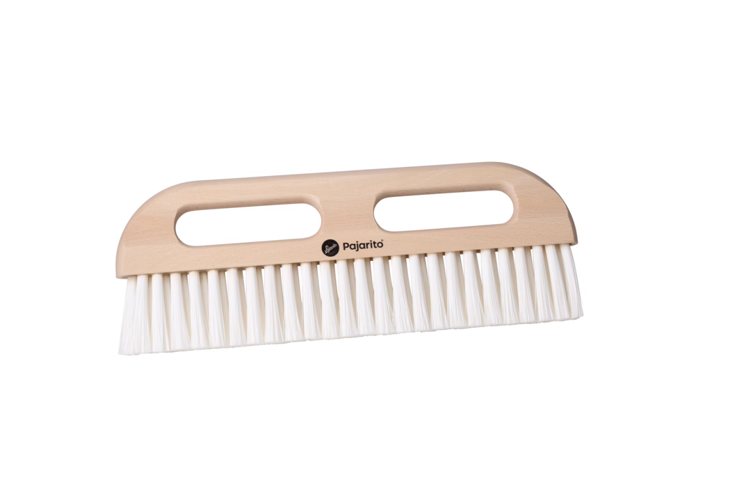 Produktaufnahme einer Holz-Handbürste mit zwei länglichen, ovalen Grifföffnungen und einem glatten, hellen Buchenholzgriff. Die Bürste besitzt dichte, gerade weiße Kunststoffborsten, die in einer durchgehenden Reihe entlang der unteren Kante sitzen. Das kleine schwarze 'Pajarito'-Logo ist mittig auf dem Griff aufgedruckt. Aufgenommen vor weißem Hintergrund mit weichem, gleichmäßigem Licht, in horizontaler Ausrichtung; geeignet zum Kehren und Abstauben im Haushalt.