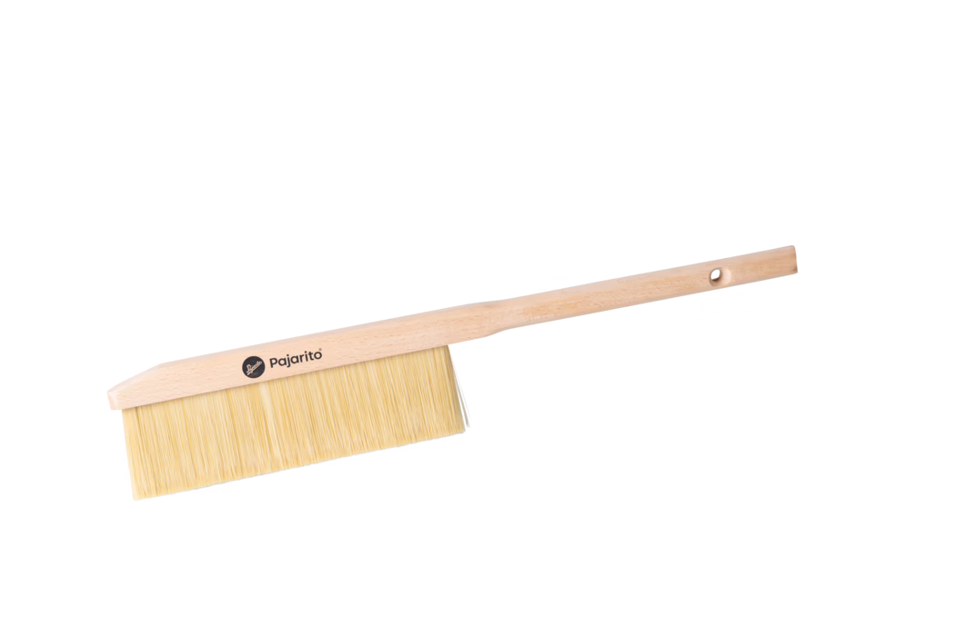 Pajarito_Dummy_Duster long handle P-Line_179114_2024-02 Nahaufnahme eines langstieligen Handbesens aus Holz vor weißem Hintergrund. Der Besen verfügt über einen hellen Buchenholzgriff und einen schlanken, rechteckigen Kopf mit dichten, hellen Naturborsten. Auf dem Holzkopf ist ein rundes Logo mit der Aufschrift „Pajarito“ aufgedruckt, und nahe dem Ende des Griffs ist ein aufgebohrtes Aufhänge-Loch sichtbar. Das schlichte, funktionale Design deutet darauf hin, dass der Besen zum Kehren oder Abstauben von Innenflächen und Werkbänken gedacht ist.