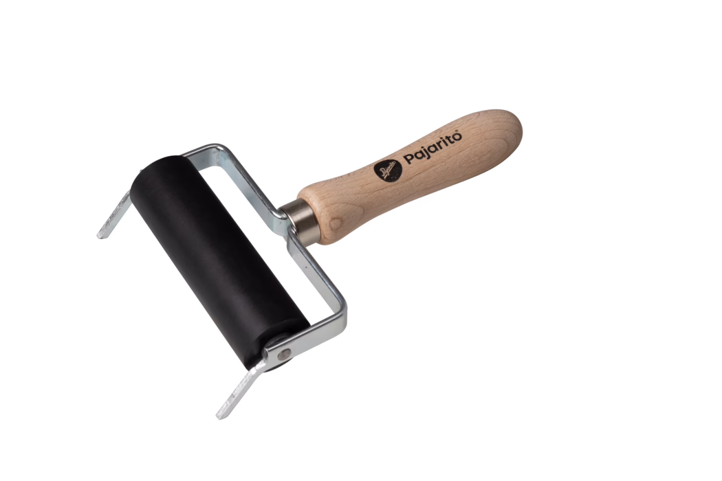 Ein kleiner Handrakel (Farbroller) auf weißem Hintergrund. Er verfügt über eine glatte schwarze Gummiwalze, die in einem gebogenen silbernen Metallgestell montiert ist, und einen ergonomisch geformten hellen Holzgriff mit dem aufgedruckten Markennamen „Pajarito“. Das Metallgestell ist mit sichtbaren Nieten an der Walze befestigt; der Holzgriff zeigt natürliche Holzmaserung und ein abgerundetes Ende. Das Werkzeug dient zum gleichmäßigen Verteilen von Druckfarbe oder Lack auf Druckplatten, Linoleum oder Papier und wirkt sauber und unbenutzt. Die Komposition ist leicht diagonal, die Walze unten links, der Griff verläuft nach oben rechts.