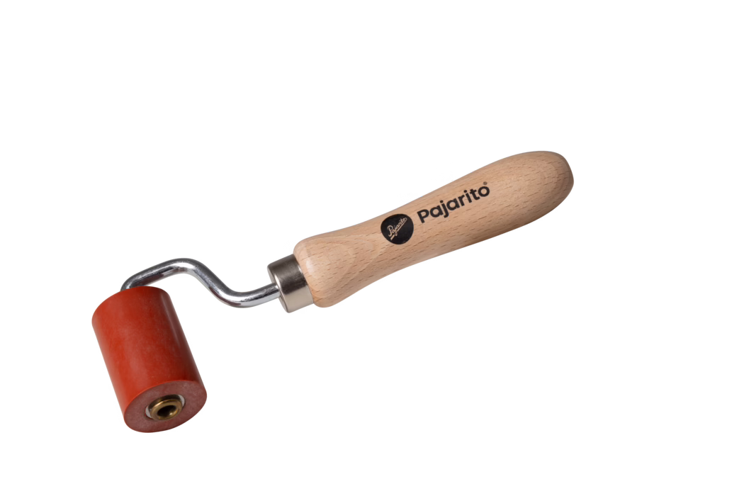 Nahaufnahme eines kleinen Handrollers vor weißem Hintergrund. Der Roller hat einen ergonomischen hellen Holzgriff mit aufgedrucktem Markennamen 'Pajarito', einen kurzen gebogenen Metallschaft mit Metallhülse und eine rote zylindrische Gummiwalze mit messingfarbener Innenbuchse. Kompaktes, handliches Werkzeug zum Glätten oder Andrücken von Materialien.