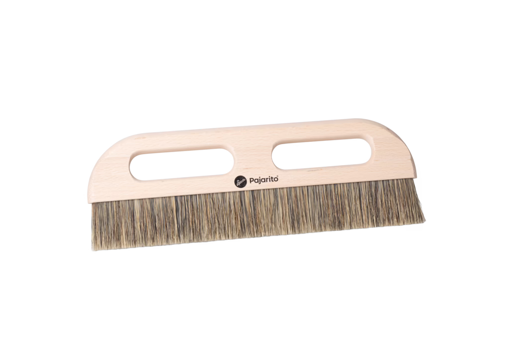 Nahaufnahme eines Holzhandbesens mit einem hellen Buchenholzgriff und dichten Naturborsten. Der flache, rechteckige Griff verfügt über zwei längliche ovale Handöffnungen für einen sicheren Zweihandhalt und ein kleines schwarzes Pajarito-Logo in der Mitte. Die Borsten bestehen aus gemischten beige- und braunfarbenen Fasern, sind gleichmäßig zu einer geraden Kante gestutzt und eignen sich zum Kehren von feinem Staub und kleinen Schmutzpartikeln von Böden oder Arbeitsflächen. Das Besenmodell ist auf einem sauberen weißen Hintergrund fotografiert und zeigt die glatt geschliffene Holzmaserung, abgerundete Ecken und ein praktisches ergonomisches Design.