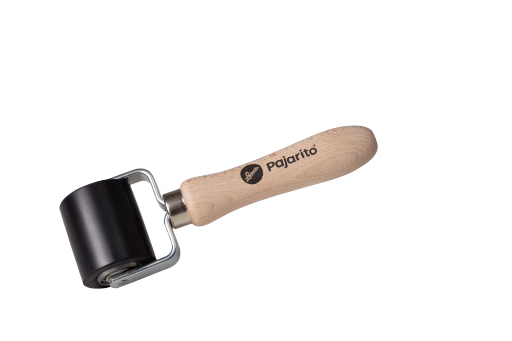 Pajarito_Dummy_Pressure roller small_038700_2021-02 Ein kleiner Handroller auf weißem Hintergrund: ein glatter schwarzer Gummizylinder auf einem Metallbügel, befestigt an einem hellen Buchenholzgriff mit dem Markenaufdruck 'pajarito'. Der ergonomisch geformte, geschliffene Griff zeigt sichtbare Holzmaserung; der Metallrahmen mit Lager ermöglicht ein freies Rollen der Walze. Das Werkzeug ist diagonal angeordnet, gut ausgeleuchtet und eignet sich zum Andrücken beim Laminieren, Glätten von Tapeten oder Folien sowie für den Druckereinsatz.
