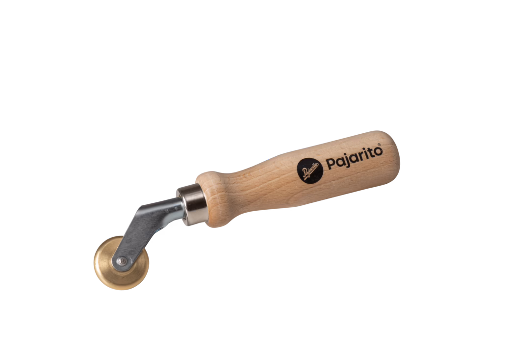 Pajarito_Dummy_Pressure roller PVC-films_034434_2021-02 Handwerkzeug mit einem glatten, abgerundeten Griff aus Buchenholz, bedruckt mit dem Markennamen 'Pajarito', einer kurzen Metallwelle und einer kleinen Messingrolle auf einem drehbaren Halter; zum Andrücken und Glätten von Nähten, Tapetenrändern, Leder oder Klebeflächen. Die natürliche Holzmaserung und das schwarze Logo sind gut sichtbar. Aufnahme vor weißem Hintergrund.
