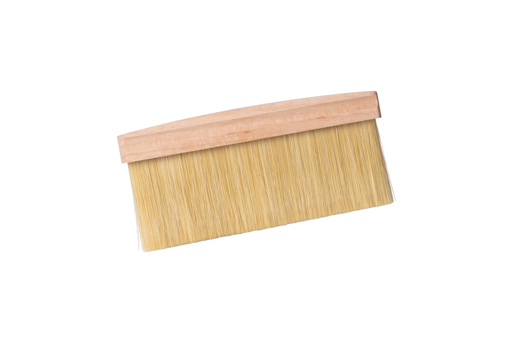 Ein breiter, flacher Pinsel mit hellem Holzgriff und dicht stehenden, hellbeigen synthetischen Borsten, auf weißem Hintergrund leicht diagonal dargestellt. Der Pinsel wird typischerweise zum Tapezieren, Abstauben oder Auftragen von Lacken verwendet; die langen, dicht angeordneten Borsten sitzen an einem einfachen, unbehandelten Holzrücken.